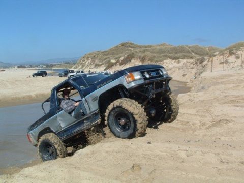 1989 Ford Bronco - A modified Ford Bronco navigates a sandy incline near the ocean, with other vehicles in the background.