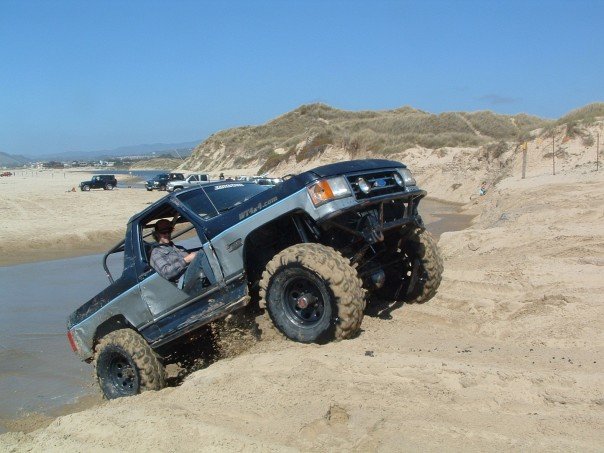 Topless blue and silver Ford Bronco climbing a wet sand wash at coastal dunes