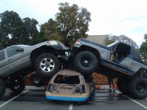 1989 Ford Bronco - Modified trucks are stacked on a wrecked car in a parking lot.