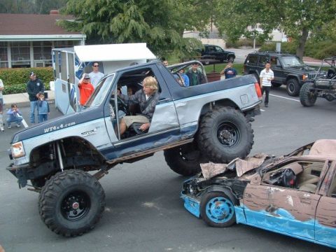 1989 Ford Bronco - A heavily modified truck with large tires is shown driving over a wrecked car, with spectators in the background.