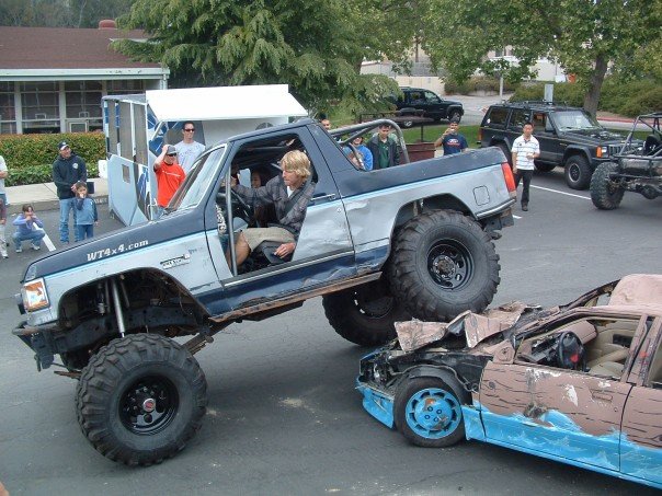 Lifted two-tone blue Ford 4x4 driving its front wheels onto a crushed blue sedan at an outdoor event