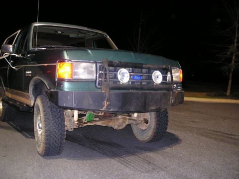 1989 Ford Bronco - A green Ford Bronco with a heavy-duty winch bumper and two round auxiliary lights is parked on asphalt at night.