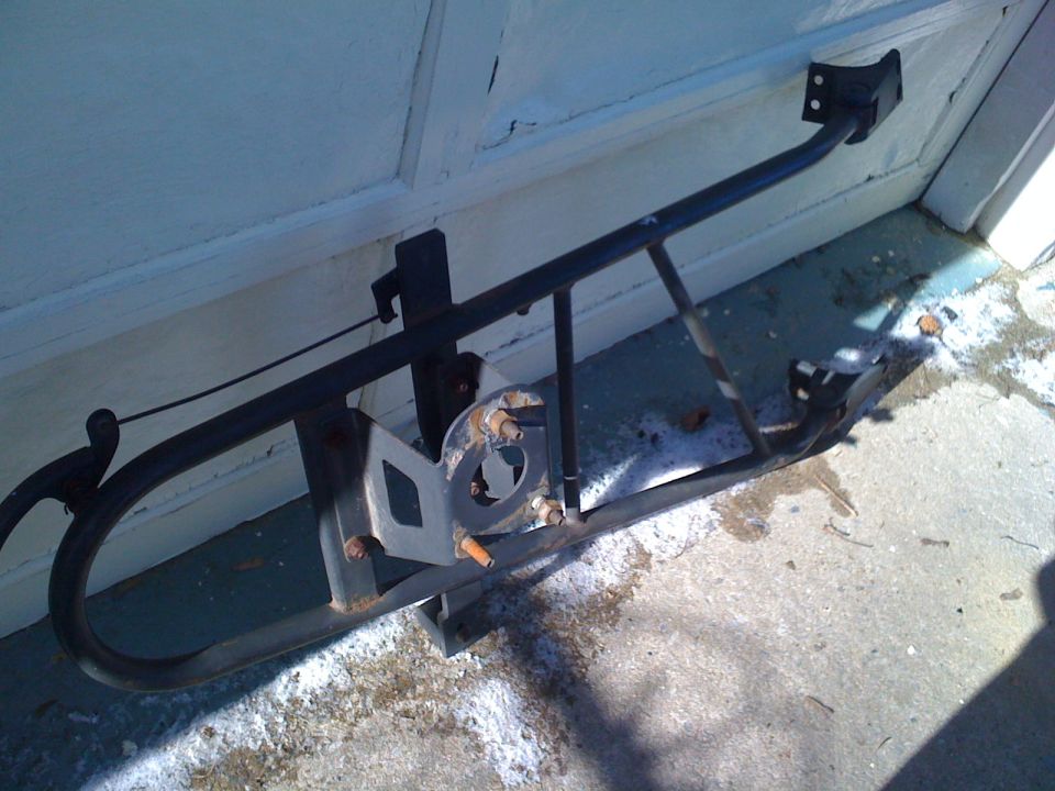 Black swing-away spare tire carrier for a Ford Bronco leaning against a light blue garage door