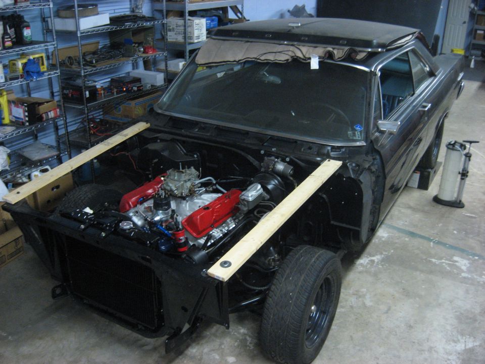 Ford hardtop with fenders off, wood jigs across frame rails and red V8 installed in shop