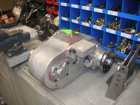 1996 Ford Bronco - A silver transfer case sits on a workbench next to a blue parts organizer filled with bolts and hardware. The transfer case has red accents…