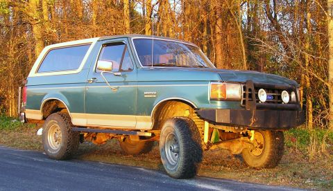 1989 Ford Bronco - Green and tan two-door Ford Bronco with a white hardtop parked on a roadside in golden late-day light, shot from the front three-quarter. I…