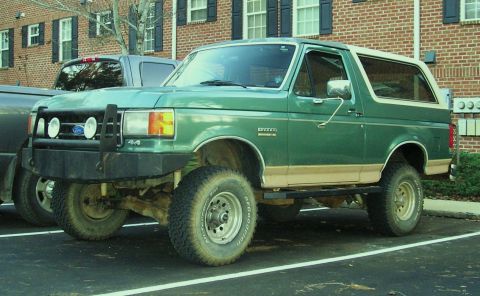 1989 Ford Bronco - Mud-splattered green full-size Ford Bronco with a tan hardtop, shot from the driver's-side front three-quarter in a parking lot beside a br…