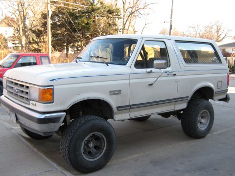 1987 Ford Bronco - This lifted Ford Bronco is parked outside on a paved surface with trees and houses in the background.