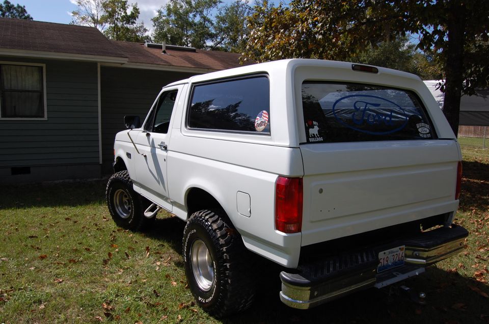 White Ford Bronco with lifted suspension and aftermarket wheels, parked in front of a house. License plate reads 519 0724