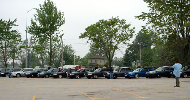 Fleet of dark sedans, one blue sedan, parked in a lot. A man in a blue shirt stands near the right