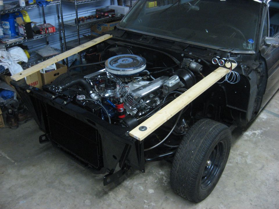 Full-size car with front sheetmetal removed, V8 installed and 2x4 boards bracing the fender aprons