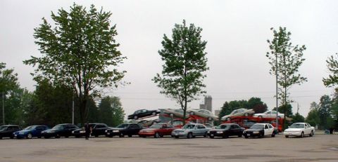 2000 Ford Crown Victoria - A collection of sedans are visible, some parked and others loaded onto transport vehicles under an overcast sky.