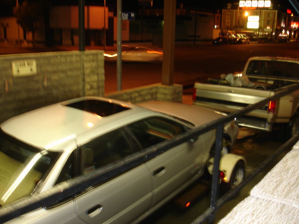 White sedan on a trailer behind a silver pickup truck at night.
