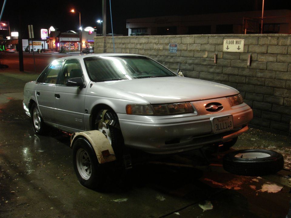 Silver Ford Taurus sedan at night with front wheel off and spare tire on ground