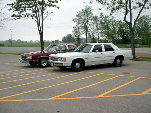 2000 Ford Crown Victoria - Two older model sedans are parked in a lot with yellow parking lines.