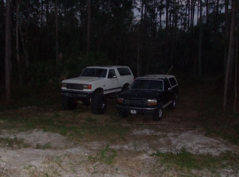 1989 Ford Bronco - Two Ford Bronco SUVs are parked in a wooded area at dusk. The white Bronco on the left is lifted with large tires, while the black Bronco o…