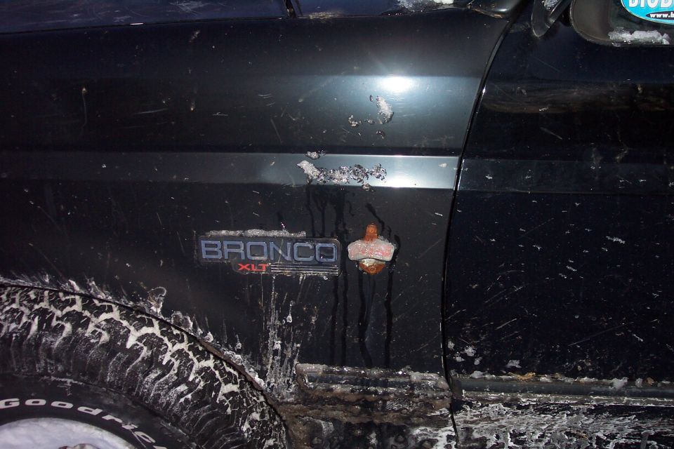Black 1992 Ford Bronco fender and door with "BRONCO XLT" emblem and bottle opener, covered in dirt and snow.