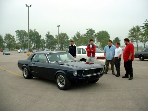 2000 Ford Crown Victoria - Several people gather around a dark blue classic Ford Mustang coupe in a parking lot.