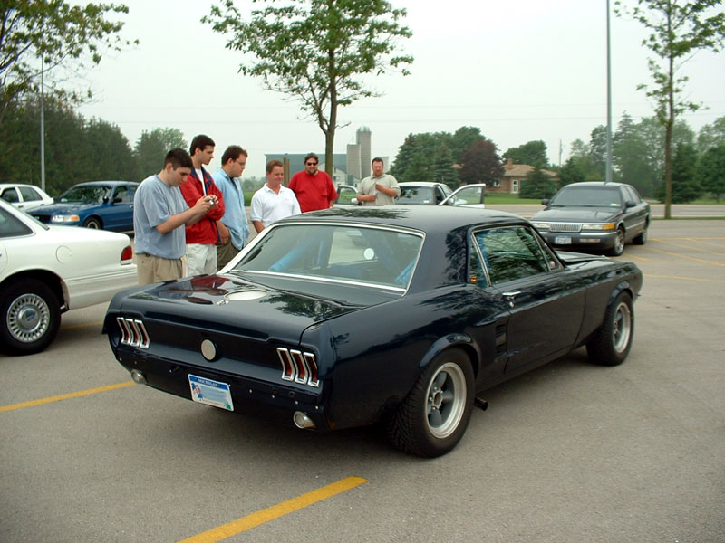 Dark 1967 Ford Mustang coupe parked as men inspect it in a lot
