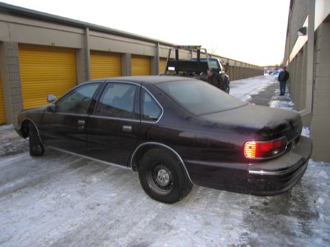 1991 Ford LTD Crown Victoria - Rear three-quarter photo of a dark 1990s Chevrolet Caprice sedan on snow-covered pavement near orange storage-unit doors.