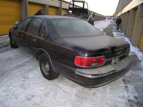 1991 Ford LTD Crown Victoria - Rear photo of a dark Chevrolet Caprice sedan with brake lights illuminated, parked beside a tow truck in a snowy storage lot.
