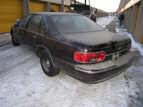 1991 Ford LTD Crown Victoria - Rear three-quarter photo of a dark Chevrolet Caprice sedan near a tow truck and orange storage-unit doors in the snow.