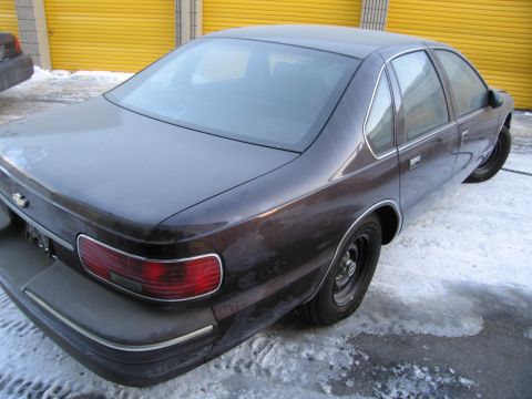 1991 Ford LTD Crown Victoria - Rear three-quarter photo of a dark Chevrolet Caprice sedan parked on snow in front of orange storage-unit doors.