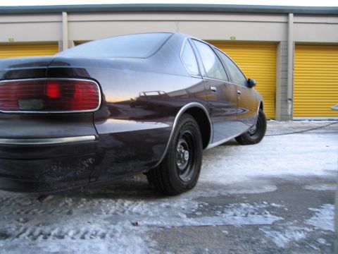 1991 Ford LTD Crown Victoria - Low-angle rear three-quarter photo of a dark Chevrolet Caprice sedan parked near storage units in the snow.