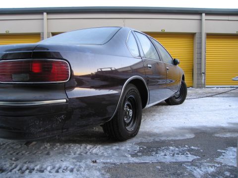 1991 Ford LTD Crown Victoria - Low-angle rear three-quarter photo of a dark Chevrolet Caprice sedan with a tow chain in a snowy storage lot.