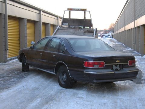 1991 Ford LTD Crown Victoria - Rear three-quarter photo of a black Chevrolet Caprice sedan parked beside a flatbed tow truck in a snowy lot.