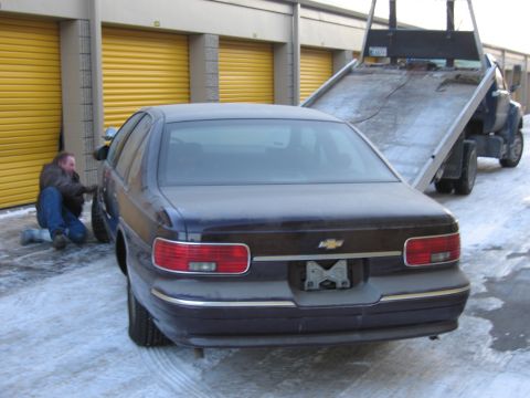 1991 Ford LTD Crown Victoria - Rear photo of a dark Chevrolet Caprice sedan with a person crouched at the front wheel beside a flatbed tow truck in snow.