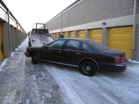 1991 Ford LTD Crown Victoria - Side view of a dark Chevrolet Caprice sedan positioned at a flatbed tow ramp in a snowy storage alley.