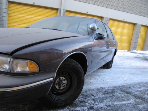 1991 Ford LTD Crown Victoria - Low-angle front three-quarter photo of a dark Chevrolet Caprice sedan parked by storage units in the snow.