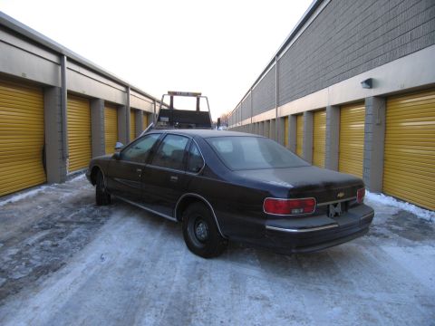 1991 Ford LTD Crown Victoria - Rear three-quarter photo of a black Chevrolet Caprice sedan parked between storage-unit rows with a tow truck behind in the snow.