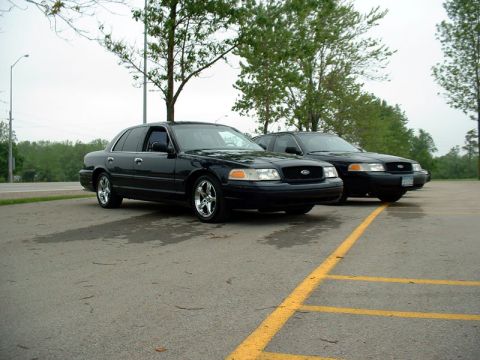 2000 Ford Crown Victoria - Two black Ford Crown Victorias are parked side-by-side in a wet parking lot under an overcast sky.