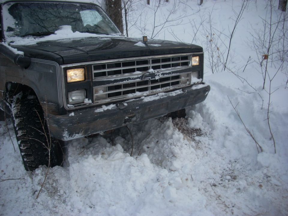 Dark Chevrolet truck stuck in snow on a wooded trail.