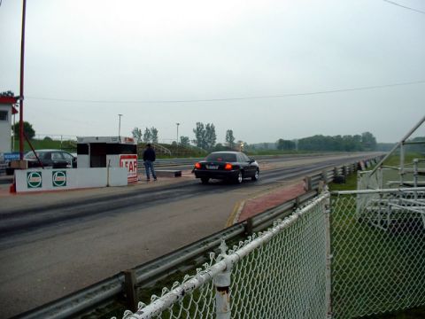 2000 Ford Crown Victoria - A black sedan speeds down the drag strip under an overcast sky, with another vehicle visible at the starting line.