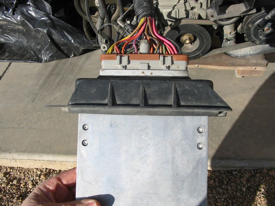 Close-up of a vehicle computer module with wires, held by a hand, in front of an engine.