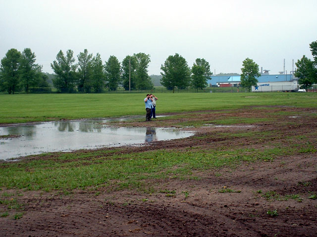 Two people beside a flooded muddy field with tire ruts and an industrial building beyond