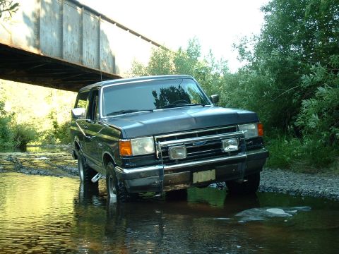 1991 Ford Bronco - A Ford Bronco navigates a stream beneath a railway bridge, surrounded by lush greenery.