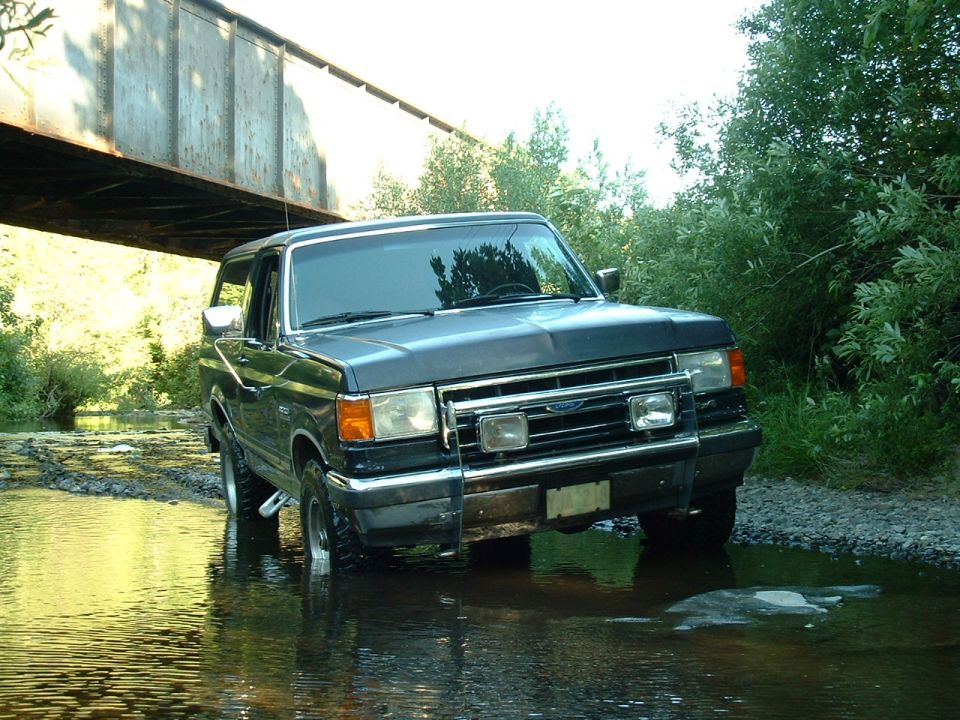 Dark blue lifted Ford Bronco with brush guard standing in a shallow creek under a steel bridge