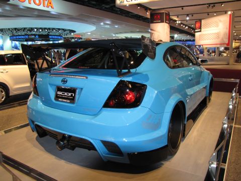 1996 Ford Probe - The rear passenger side of a bright blue Scion tC with a large rear spoiler and wide body kit is displayed at an auto show. The car is on a…