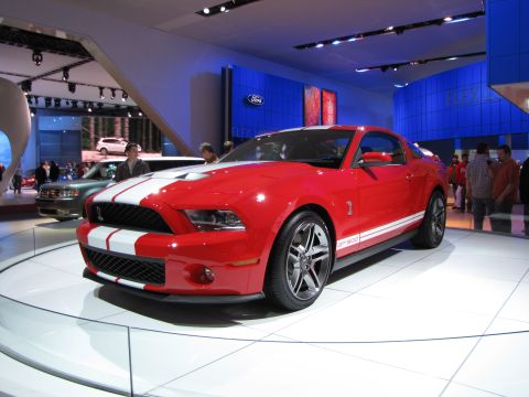 1996 Ford Probe - A bright red Ford Mustang GT500 with white racing stripes is displayed on a white platform at an auto show. The car is shown from a three-q…