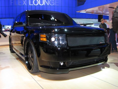 1996 Ford Probe - A black custom Ford Flex is displayed at an auto show with a large mesh grille and custom wheels. The background features a blue wall with…