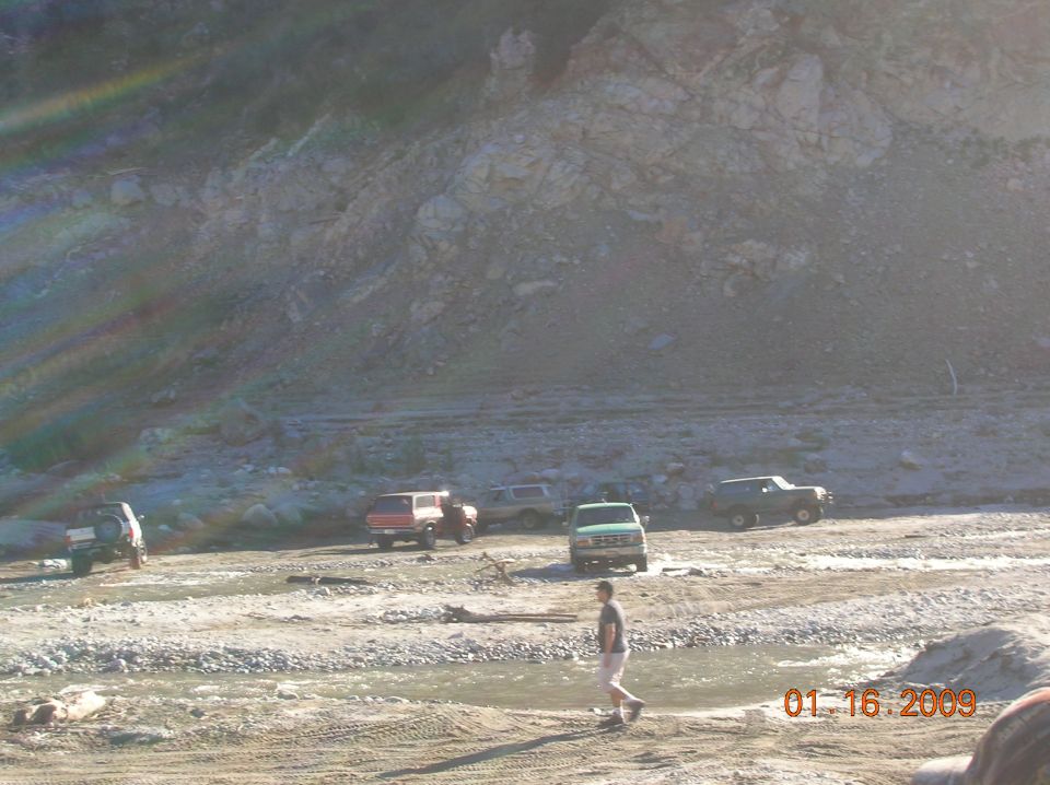 Several off-road vehicles parked in a dry riverbed with a man walking.
