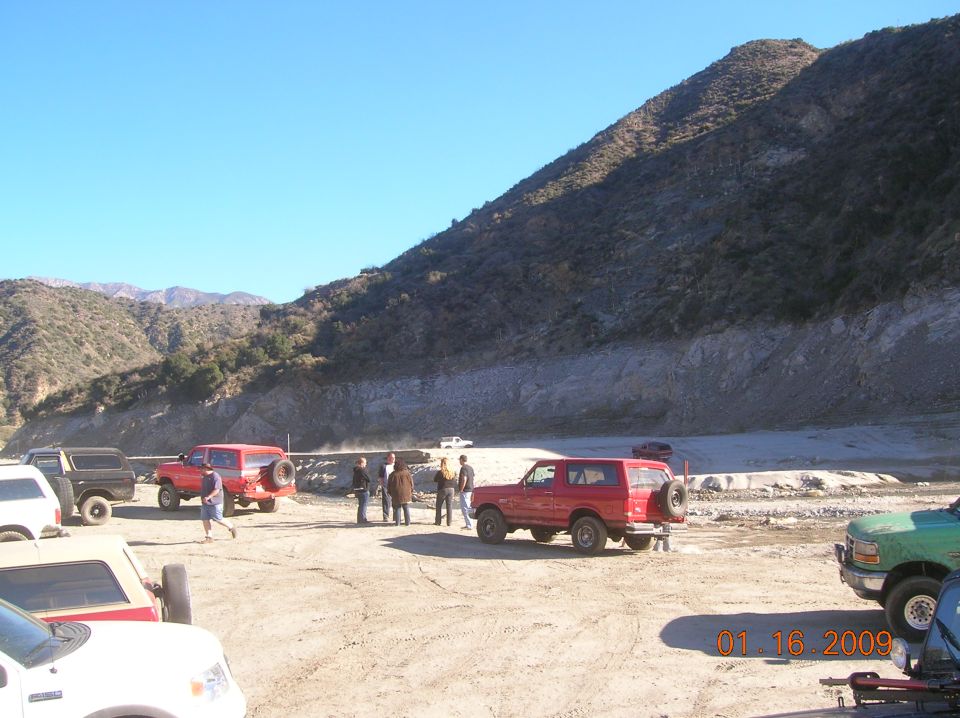 Several red Ford Broncos parked in a dirt lot with a rocky hillside and blue sky.