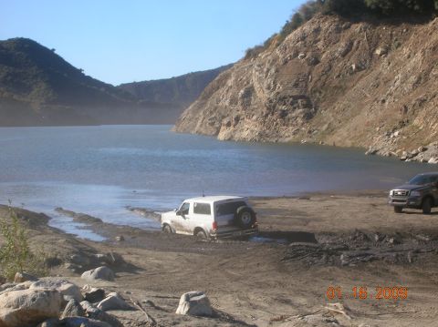 1985 Ford Bronco - A white SUV is stuck in the mud near the edge of a body of water, with another vehicle visible in the background.