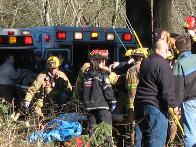 Emergency responders near an ambulance with a stretcher in a wooded area.