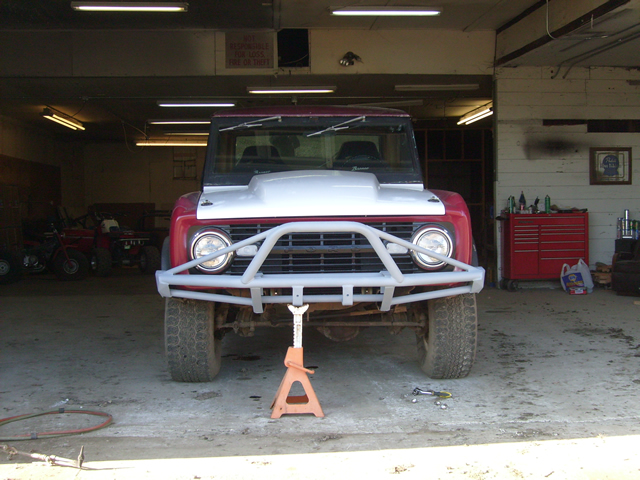 Red and white Ford Bronco with custom bumper in a garage, supported by a jack stand.