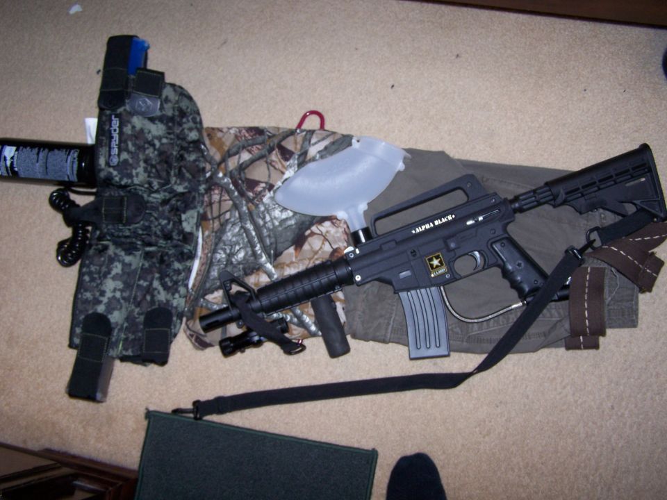Black tactical paintball or airsoft rifle laid on camo gear on carpet